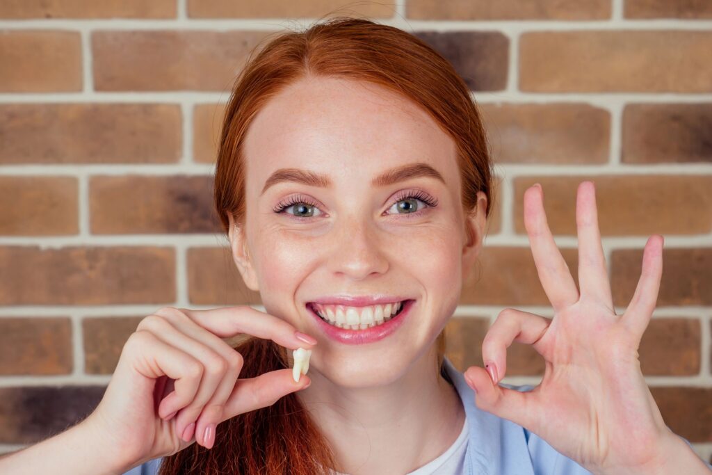 Woman with red hair holding extracted tooth in one hand and making "ok" sign with other